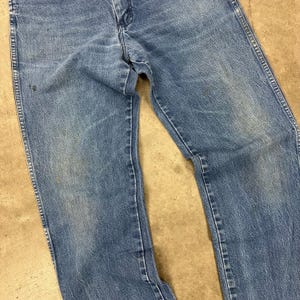 K&ouml;nnte beinhalten: Nahaufnahme einer verblichenen blauen Jeans. Die Jeans weist Gebrauchsspuren auf, mit sichtbarem Ausbleichen und Verf&auml;rbungen. Die N&auml;hte sind intakt, und die Jeans scheint ein klassischer, gerader Schnitt zu sein. Der Stoff hat eine leicht abgenutzte Textur.