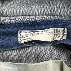 K&ouml;nnte beinhalten: Nahaufnahme einer Jeans aus Denim mit einem sichtbaren Etikett. Das Etikett lautet "Wrangler" und gibt die Gr&ouml;&szlig;e 31 x 34 Zoll (78,7 x 86,4 cm) an. Das Etikett enth&auml;lt auch Waschanweisungen und die Aufschrift "Made in USA" und "100% Baumwolle."