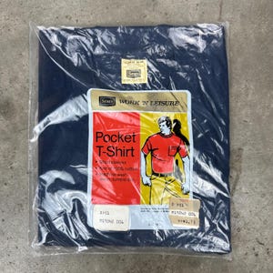 May include: A navy blue Pocket T-Shirt from Sears, still in its original packaging. The label reads "Work 'N Leisure" and features an illustration of a man wearing a red pocket t-shirt. The shirt is knit of 100% cotton.