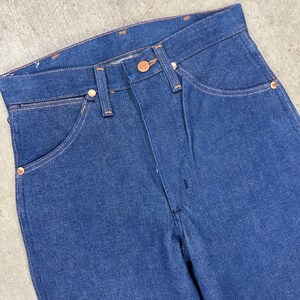 May include: Close-up of a pair of blue denim jeans. The jeans have a classic five-pocket design with copper-colored rivets and a button closure. The stitching is a contrasting orange color.
