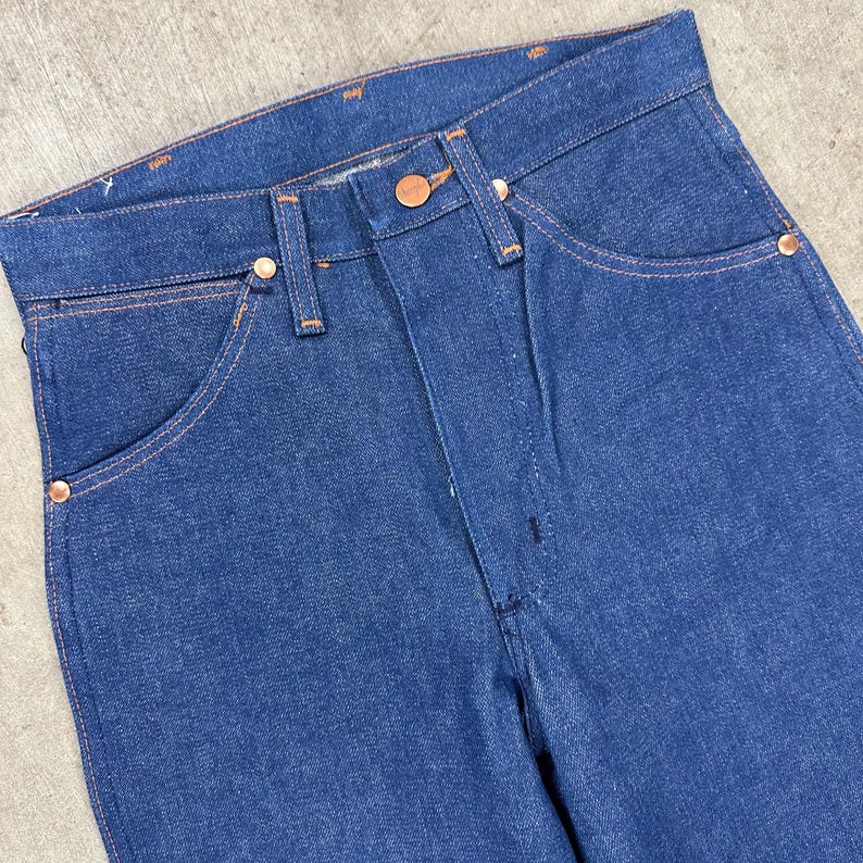 May include: Close-up of a pair of blue denim jeans. The jeans have a classic five-pocket design with copper-colored rivets and a button closure. The stitching is a contrasting orange color.