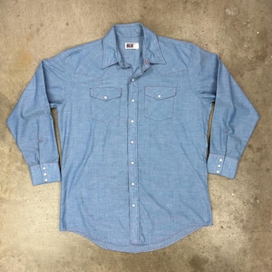 May include: Light blue long-sleeve button-down shirt with two chest pockets and white buttons. The shirt has red stitching details and a classic Western style.