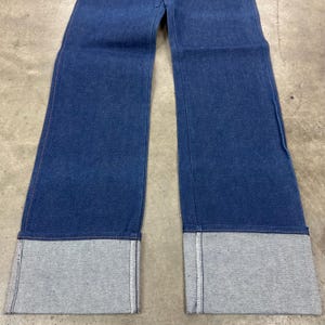 May include: Blue denim jeans with wide legs and large, gray cuffs. The jeans are made of a medium-weight denim fabric and have a classic five-pocket design. The cuffs are approximately 3 inches (7.6 cm) wide.