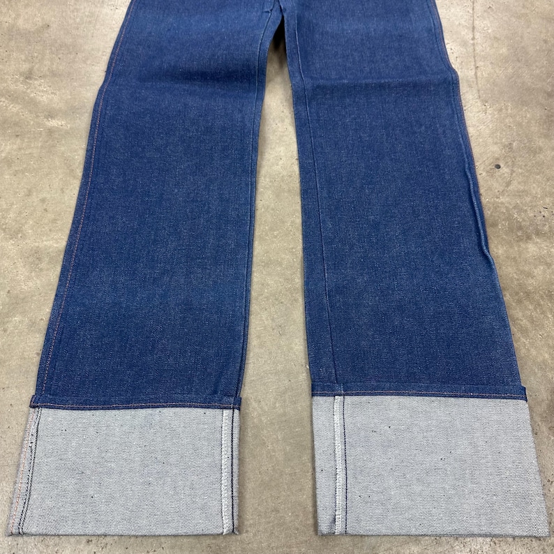 May include: Blue denim jeans with wide legs and large, gray cuffs. The jeans are made of a medium-weight denim fabric and have a classic five-pocket design. The cuffs are approximately 3 inches (7.6 cm) wide.