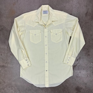 May include: Pale yellow long-sleeve shirt with a Western-style design. Features include a pointed collar, two chest pockets, and snap closures. The shirt has a curved hem and buttoned cuffs. A small tag is visible near the collar.