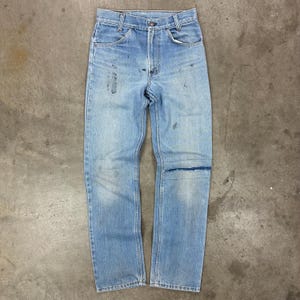 May include: Light blue denim jeans with a classic five-pocket design. The jeans show signs of wear, including faded areas and a knee patch. The jeans are laid flat on a concrete surface.