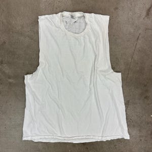 May include: A white sleeveless t-shirt with a crew neck. The shirt has been cut at the sleeves, creating a muscle shirt style. The fabric appears to be a lightweight cotton, and the shirt is slightly wrinkled.