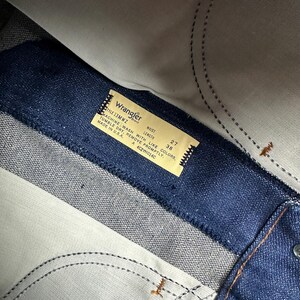 May include: Close-up of a pair of blue denim jeans with a Wrangler brand tag. The tag indicates a waist size of 27 inches and a length of 38 inches. The jeans are made in the U.S.A.
