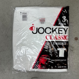 May include: A package of three white Jockey Classic T-shirts. The packaging is clear plastic with red and black accents. The Jockey logo is visible, along with the text "3 Pack" and "100% Combed Cotton."