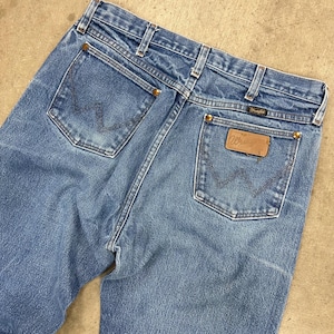 May include: Light blue denim jeans with the Wrangler logo on the back pocket. The jeans have a classic five-pocket design with brass rivets and a faded wash. The back pockets feature the signature Wrangler stitching.