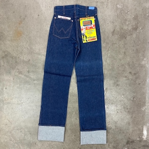 May include: Dark blue denim jeans with a straight leg cut and folded cuffs. The jeans have a high waist and a Wrangler label on the back pocket. A yellow and red tag with the Wrangler logo is attached.