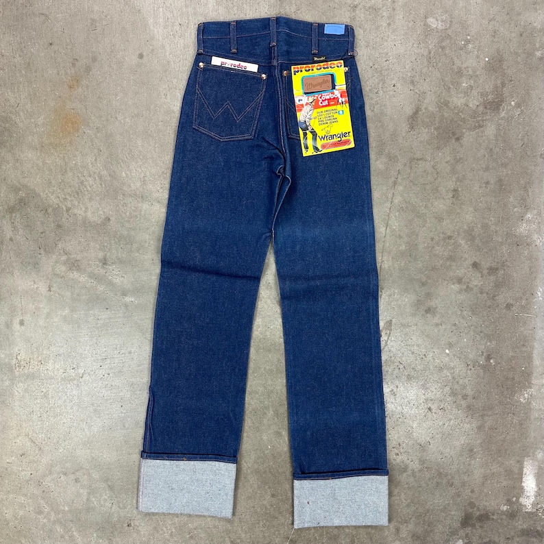 May include: Dark blue denim jeans with a straight leg cut and folded cuffs. The jeans have a high waist and a Wrangler label on the back pocket. A yellow and red tag with the Wrangler logo is attached.