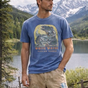 May include: Blue short-sleeved t-shirt featuring a fish graphic and the words "WHITE WATER FISHING TACKLE" in orange. The shirt is paired with khaki shorts and a cap, set against a mountain and lake landscape.