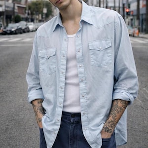 May include: Light blue button-down shirt with two chest pockets and pearl snap closures. The shirt is worn over a white tank top and paired with dark blue jeans. The sleeves are rolled up.