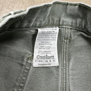 May include: Close-up of olive green Carhartt work pants. The tag reads "Carhartt" and includes the size 34 x 34. The tag also states the pants are 100% cotton and made in Mexico with U.S.A. components.