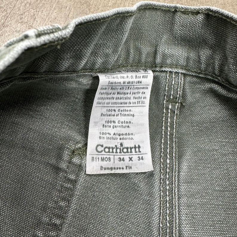 May include: Close-up of olive green Carhartt work pants. The tag reads "Carhartt" and includes the size 34 x 34. The tag also states the pants are 100% cotton and made in Mexico with U.S.A. components.