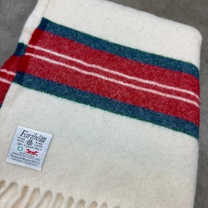 May include: Cream-colored wool blanket with red and teal stripes and white accents. The blanket has a fringed edge and a tag that reads "Faribo" and "Made in USA".