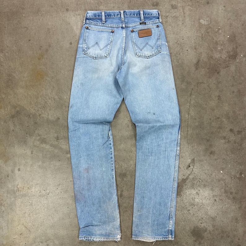 Op de afbeelding: Lichtblauwe denim jeans met een klassiek ontwerp met vijf zakken. De jeans hebben een bruine leren patch op de achterkant en het Wrangler-logo. De jeans zijn vintage en vertonen gebruikssporen.