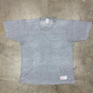 May include: A heather gray short-sleeved t-shirt with a chest pocket. The shirt has a crew neck and a small tag near the collar. A rectangular tag is sewn on the bottom right. The shirt is laid flat on a concrete surface.