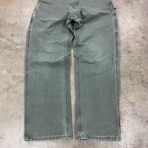 May include: A pair of olive green work pants with a straight leg design. The pants have a classic five-pocket style, a button and zipper closure, and reinforced stitching along the seams. The fabric appears to be a durable, heavy-duty material.