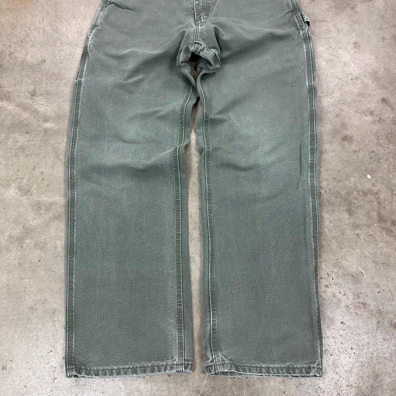 May include: A pair of olive green work pants with a straight leg design. The pants have a classic five-pocket style, a button and zipper closure, and reinforced stitching along the seams. The fabric appears to be a durable, heavy-duty material.