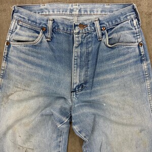 Op de afbeelding: Lichtblauwe denim jeans met een vervaagde wassing en zichtbare slijtage. De jeans heeft een klassieke vijf-zakken-stijl, een knoop- en ritssluiting en koperkleurige klinknagels. De stof vertoont tekenen van ouderdom en gebruik.