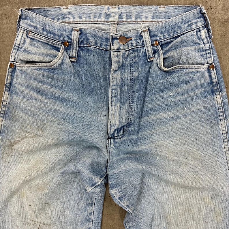 Op de afbeelding: Lichtblauwe denim jeans met een vervaagde wassing en zichtbare slijtage. De jeans heeft een klassieke vijf-zakken-stijl, een knoop- en ritssluiting en koperkleurige klinknagels. De stof vertoont tekenen van ouderdom en gebruik.
