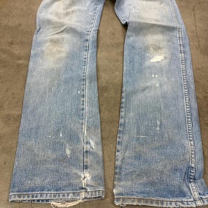 Op de afbeelding: Lichtblauwe denim jeans met distressed details, waaronder verfspatten en versleten plekken. De jeans hebben een klassiek ontwerp met vijf zakken en een licht uitlopende pijpopening. De stof vertoont tekenen van slijtage.