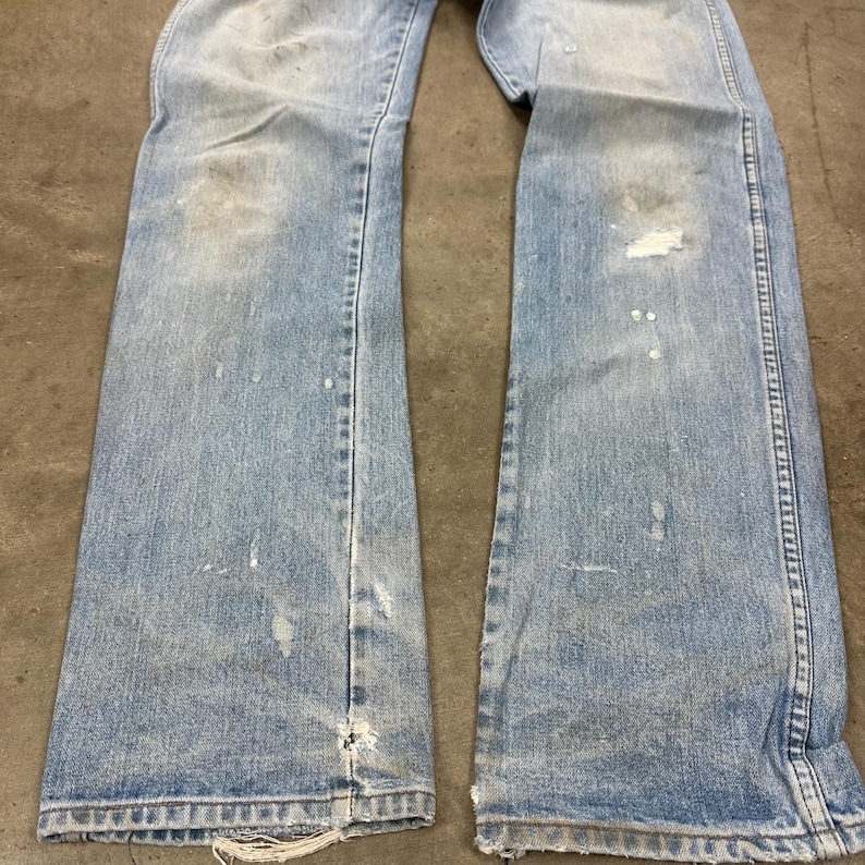 Op de afbeelding: Lichtblauwe denim jeans met distressed details, waaronder verfspatten en versleten plekken. De jeans hebben een klassiek ontwerp met vijf zakken en een licht uitlopende pijpopening. De stof vertoont tekenen van slijtage.