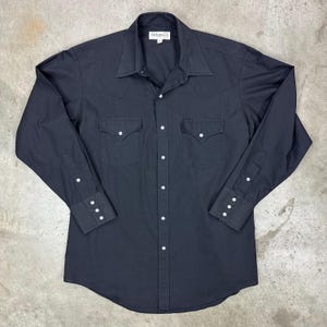May include: Black long-sleeve shirt with a Western-style design. Features a pointed collar, two chest pockets, and white button closures. The shirt has a classic, tailored fit.