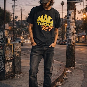 Könnte beinhalten: Eine Person trägt ein schwarzes T-Shirt mit dem Text "MAC TOOLS Racing" in Gelb und Rot. Die Person trägt auch eine dunkelblaue Jeans und eine Baseballkappe. Der Hintergrund zeigt eine Stadtstraße mit Sonnenuntergang.