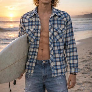 Może przedstawiać: Niebiesko-biała koszula w kratę z klasycznym kołnierzykiem i dwiema kieszeniami na piersi. Koszula jest noszona rozpięta na niebieskich dżinsowych szortach. Deska surfingowa trzymana jest w lewym ramieniu.