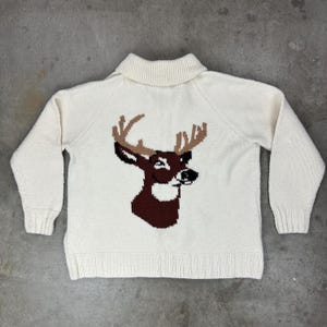 May include: Cream-colored knit sweater featuring a deer design on the front. The deer is depicted in shades of brown and black, with a detailed antler design. The sweater has a ribbed collar, cuffs, and hem, and long sleeves.