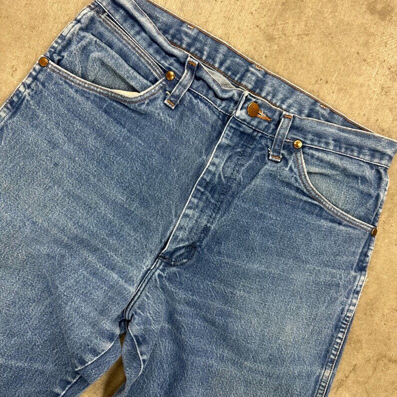 K&ouml;nnte beinhalten: Nahaufnahme einer blauen Jeans mit klassischem F&uuml;nf-Taschen-Design. Die Jeans hat einen Knopf- und Rei&szlig;verschluss sowie kupferfarbene Nieten. Der Denim hat ein verwaschenes Aussehen.