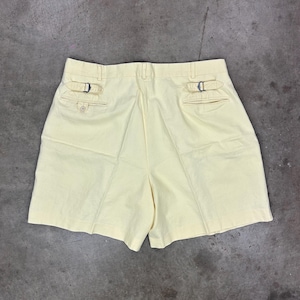 May include: Pale yellow shorts with two back pockets and adjustable side buckles. The shorts have a classic design and are made of a light-colored fabric. The shorts are a casual style.