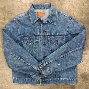 May include: A light blue denim jacket with a classic design. Features include two chest pockets, button closures, and a collar. The jacket has a worn appearance, with a small red tag on the chest pocket.