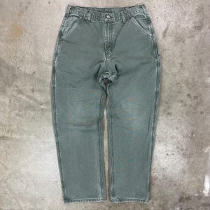 May include: A pair of sage green work pants with a button and zipper closure. The pants have multiple pockets, including a tool pocket on the right leg. The pants are made of a durable, heavy-duty fabric.