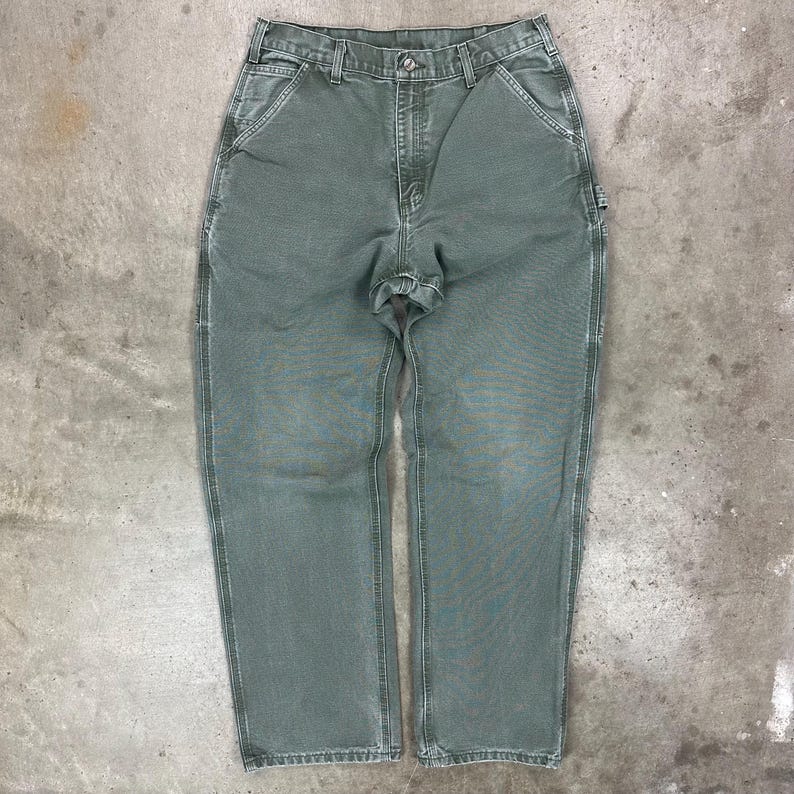 May include: A pair of sage green work pants with a button and zipper closure. The pants have multiple pockets, including a tool pocket on the right leg. The pants are made of a durable, heavy-duty fabric.