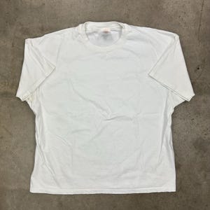 May include: A plain white t-shirt with a crew neck and short sleeves. The shirt is made of a soft, slightly wrinkled fabric. The t-shirt is laid flat on a neutral surface.