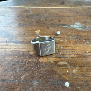 May include: A silver signet ring with a square face. The face of the ring features an engraved stylized letter "S". The ring is set on a wooden surface.
