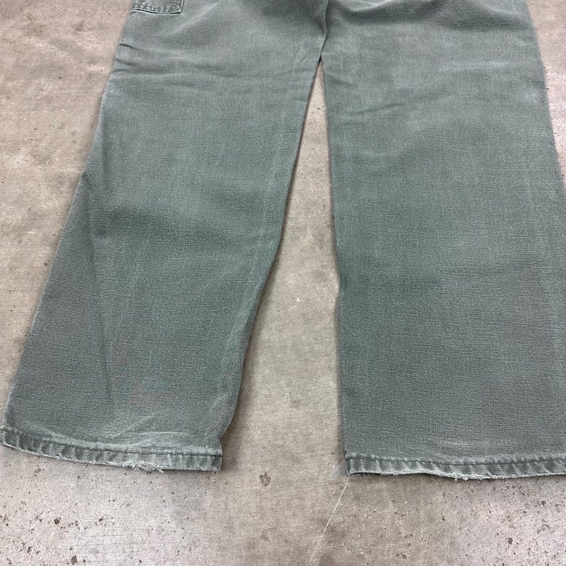 May include: A pair of faded green denim pants. The pants have a classic five-pocket design, with visible wear and tear around the edges. The fabric appears soft and well-worn, suggesting comfort and durability. The pants are a muted green color.