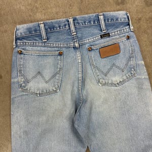 Op de afbeelding: Lichtblauwe spijkerbroek met het Wrangler-logo op de achterzak en een bruine leren patch. De jeans heeft een klassieke vijf-zakken-stijl, riemlussen en een vervaagde wassing. De achterzakken hebben de kenmerkende 'W'-stiksels van Wrangler.