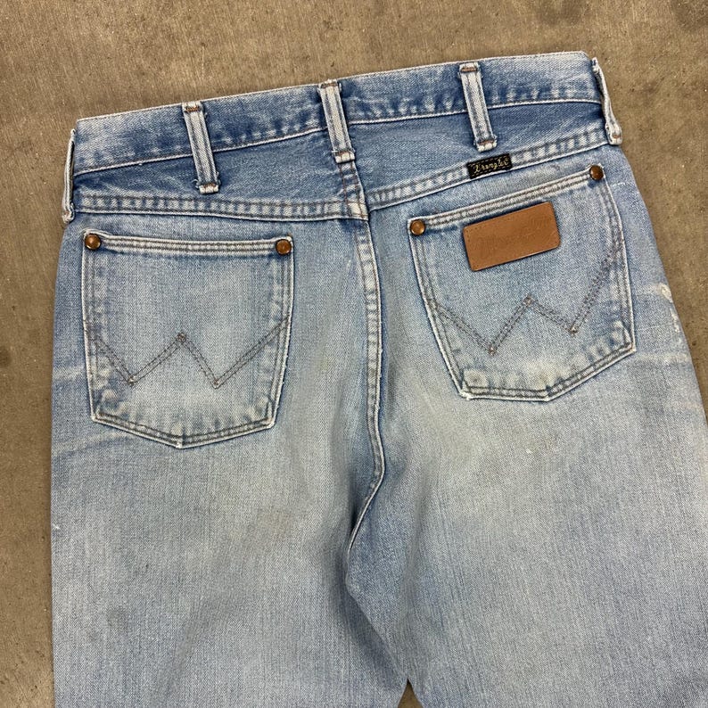 Op de afbeelding: Lichtblauwe spijkerbroek met het Wrangler-logo op de achterzak en een bruine leren patch. De jeans heeft een klassieke vijf-zakken-stijl, riemlussen en een vervaagde wassing. De achterzakken hebben de kenmerkende 'W'-stiksels van Wrangler.