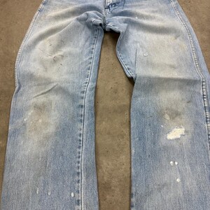 Op de afbeelding: Lichtblauwe denim jeans met zichtbare slijtage, waaronder vervaagde plekken, verfspatten en een kleine scheur. De jeans liggen plat, waarbij de voorkant van het kledingstuk te zien is. De algehele uitstraling suggereert een vintage- of workwear-stijl.