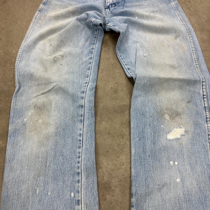 Op de afbeelding: Lichtblauwe denim jeans met zichtbare slijtage, waaronder vervaagde plekken, verfspatten en een kleine scheur. De jeans liggen plat, waarbij de voorkant van het kledingstuk te zien is. De algehele uitstraling suggereert een vintage- of workwear-stijl.