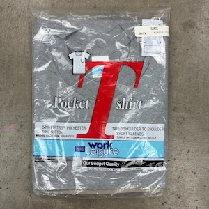 May include: A gray pocket shirt in a plastic package. The shirt has short sleeves and is made of a blend of polyester and cotton. The packaging includes the text "Pocket shirt" and "Work Leisure".