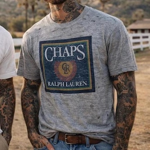 May include: A grey Chaps Ralph Lauren t-shirt with a distressed appearance. The shirt displays a rectangular graphic with the brand name and logo in navy, gold, and green. The model is wearing blue jeans and a brown belt.