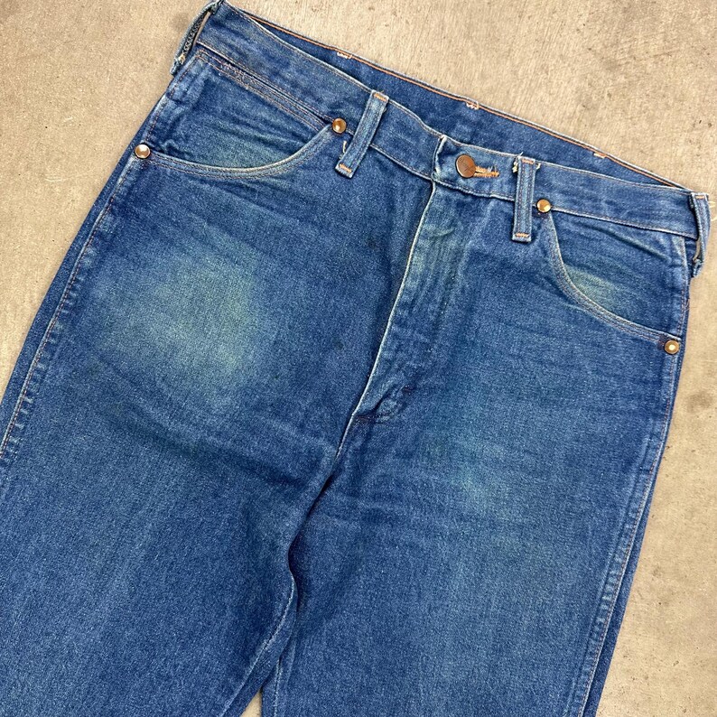 K&ouml;nnte beinhalten: Nahaufnahme einer blauen Jeans. Die Jeans hat ein klassisches F&uuml;nf-Taschen-Design mit kupferfarbenen Nieten und einem Knopfverschluss. Der Denim hat ein verwaschenes Aussehen mit helleren Bereichen auf der Vorderseite.
