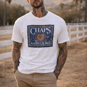 May include: White t-shirt with a rectangular graphic featuring the word "CHAPS" in white, a red and gold emblem, and "RALPH LAUREN" in white. The shirt is paired with khaki trousers.