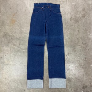May include: Dark blue denim jeans with a high waist and straight leg. The jeans have a button and zipper closure, front and back pockets, and a gray cuff at the hem. The jeans are laid flat on a concrete surface.